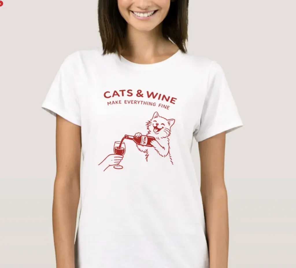 A woman wearing a white t-shirt with a red graphic illustration of a cat pouring wine into a glass held by a person, with the text "CATS & WINE MAKE EVERYTHING FINE.