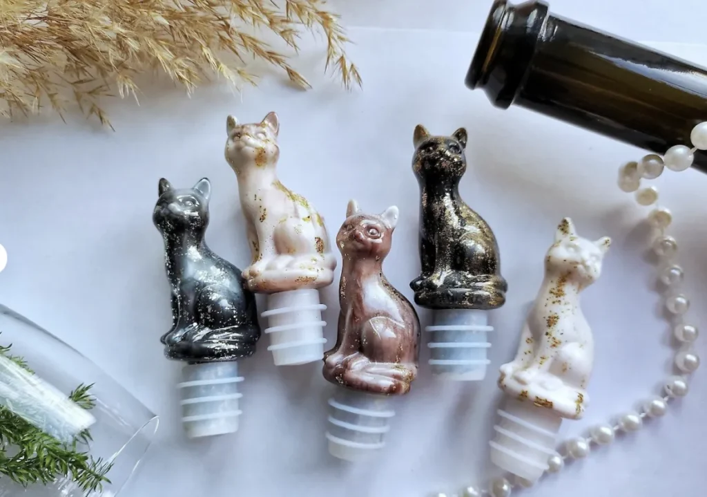 A group of five handmade artisanal cat-shaped wine bottle stoppers made of shimmering epoxy resin in various colors like black, white, and rose gold with gold leaf flakes, shown next to a wine bottle and a string of pearls.