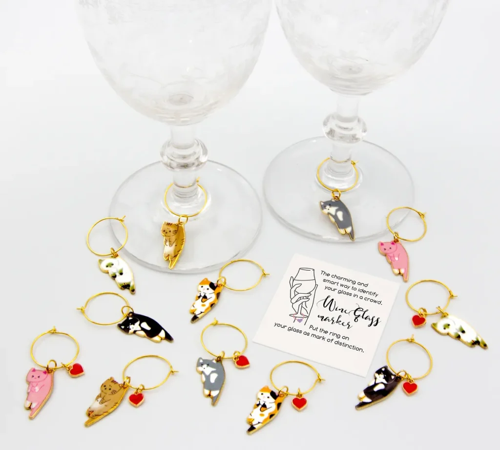 A collection of enamel cat wine charms with gold rings and small red heart pendants, shown scattered around the base of two elegant wine glasses.