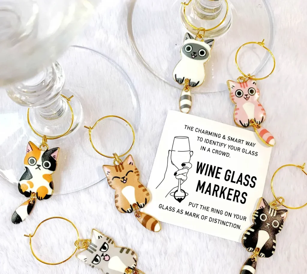 A set of six colorful cat-shaped wine glass markers with gold rings, including various patterns like calico, tabby, and siamese, displayed next to a wine glass on a white fluffy surface.